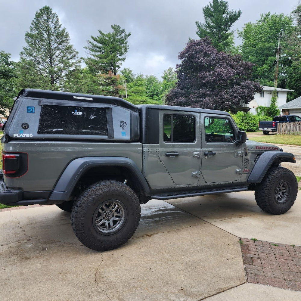 A dark gray lifted Jeep Gladiator pickup truck with a custom black canopy cover, parked on a concrete driveway in a residential backyard setting.