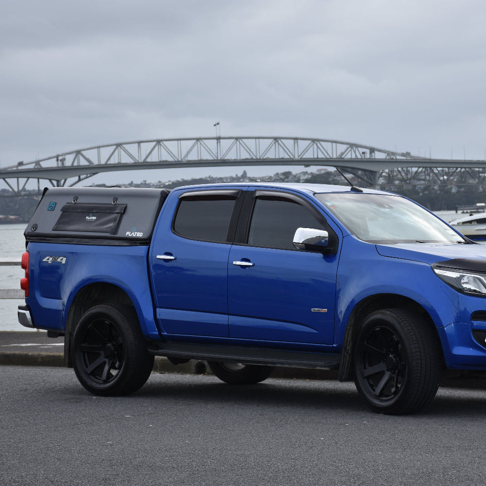 A blue pickup truck with a black hardtop canopy parked outdoors near a waterfront, with a bridge and watercraft visible in the background under an overcast sky.