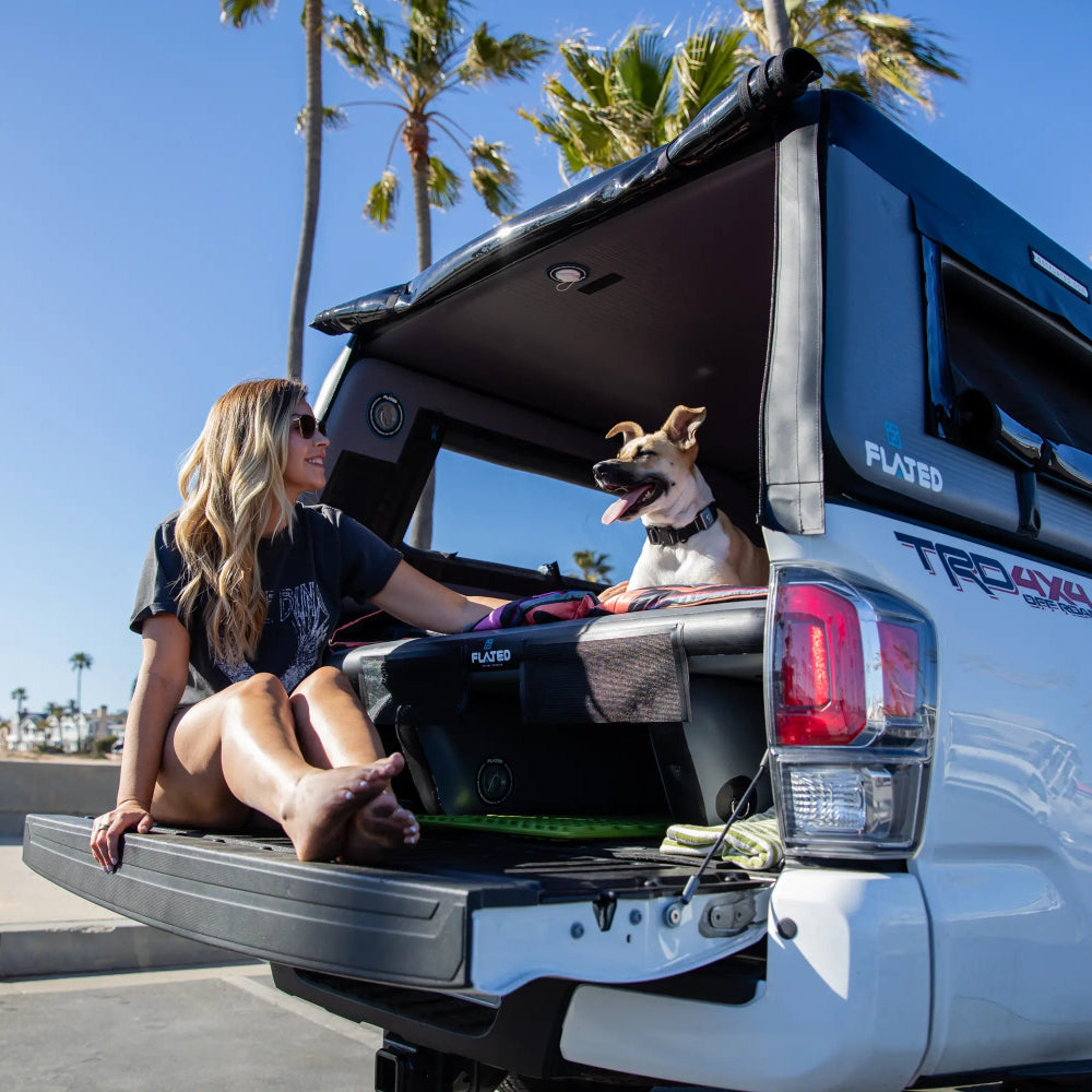 A woman sits in the open tailgate of a white pickup truck while a dog rests on the tailgate lid. The truck is equipped with a black roof-mounted canopy and a bed.